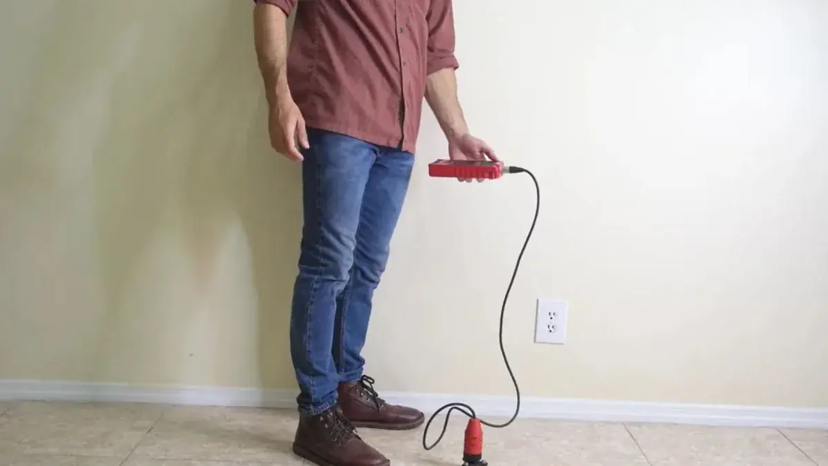 A man in jeans and boots holds a red handheld listening device connected by a cable to a sensor resting on a tile floor, in front of a plain painted wall.