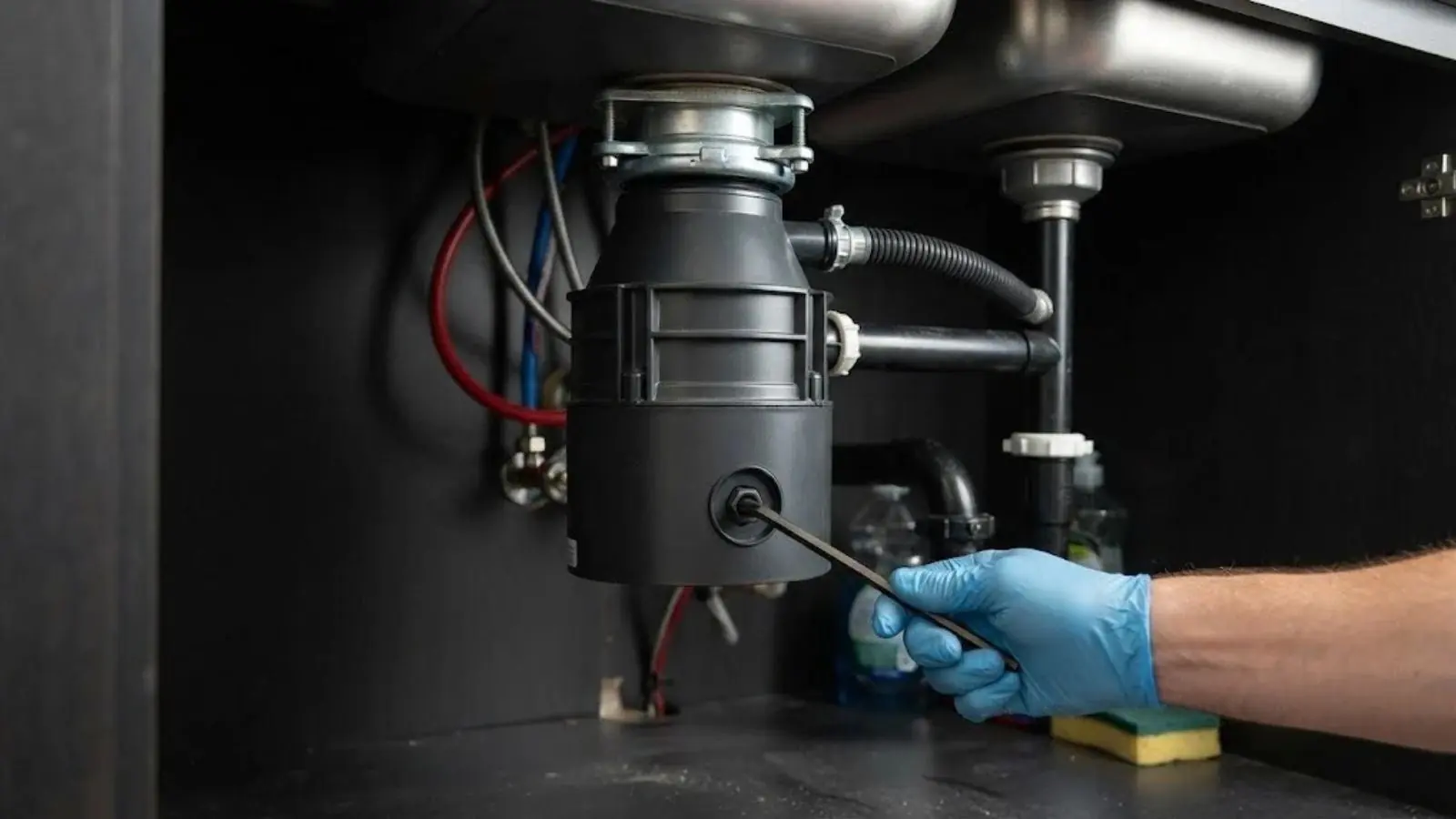 A gloved hand turns a hex key in the bottom socket of a black garbage disposal mounted under a double-bowl stainless sink, with a drain hose, P-trap, and supply lines visible beside it.
