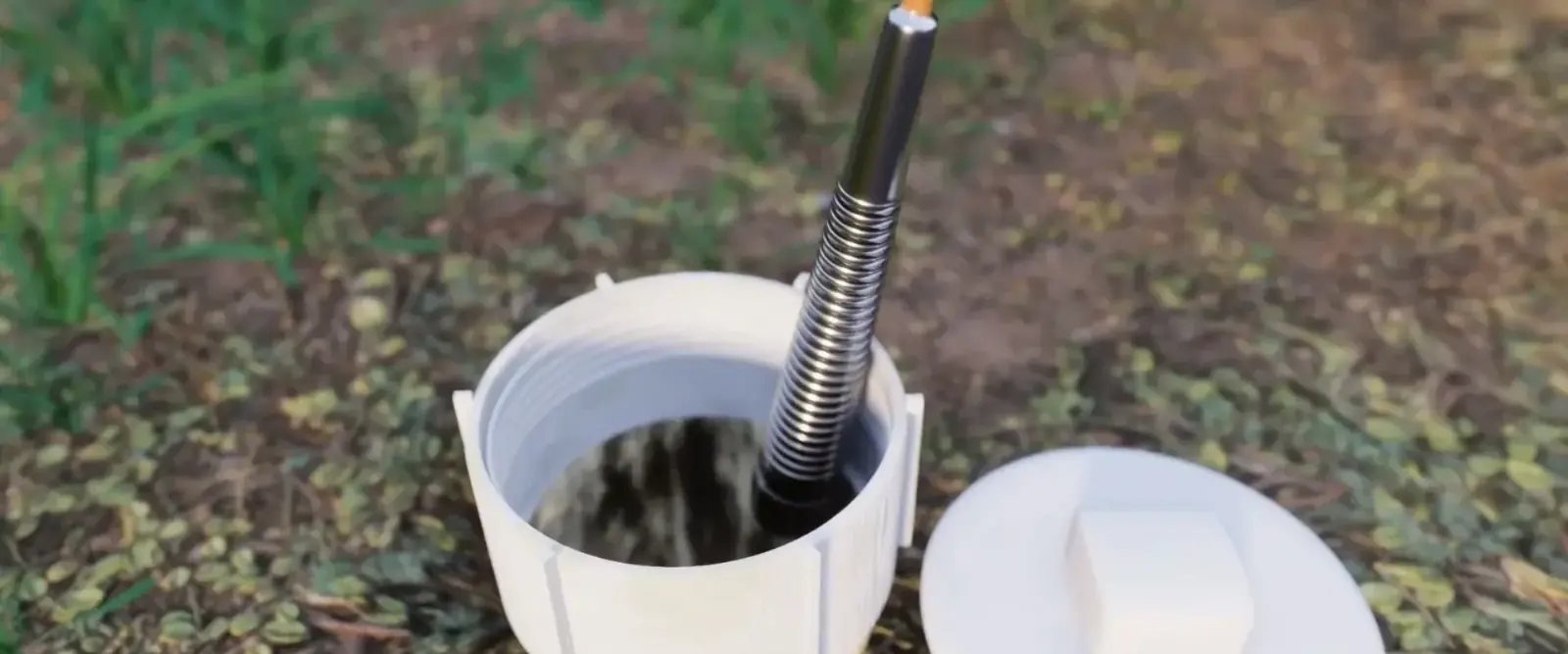A coiled steel camera cable with a bright tip is fed into an open white cleanout pipe set in grass and leaf litter, with the screw cap lying beside it.