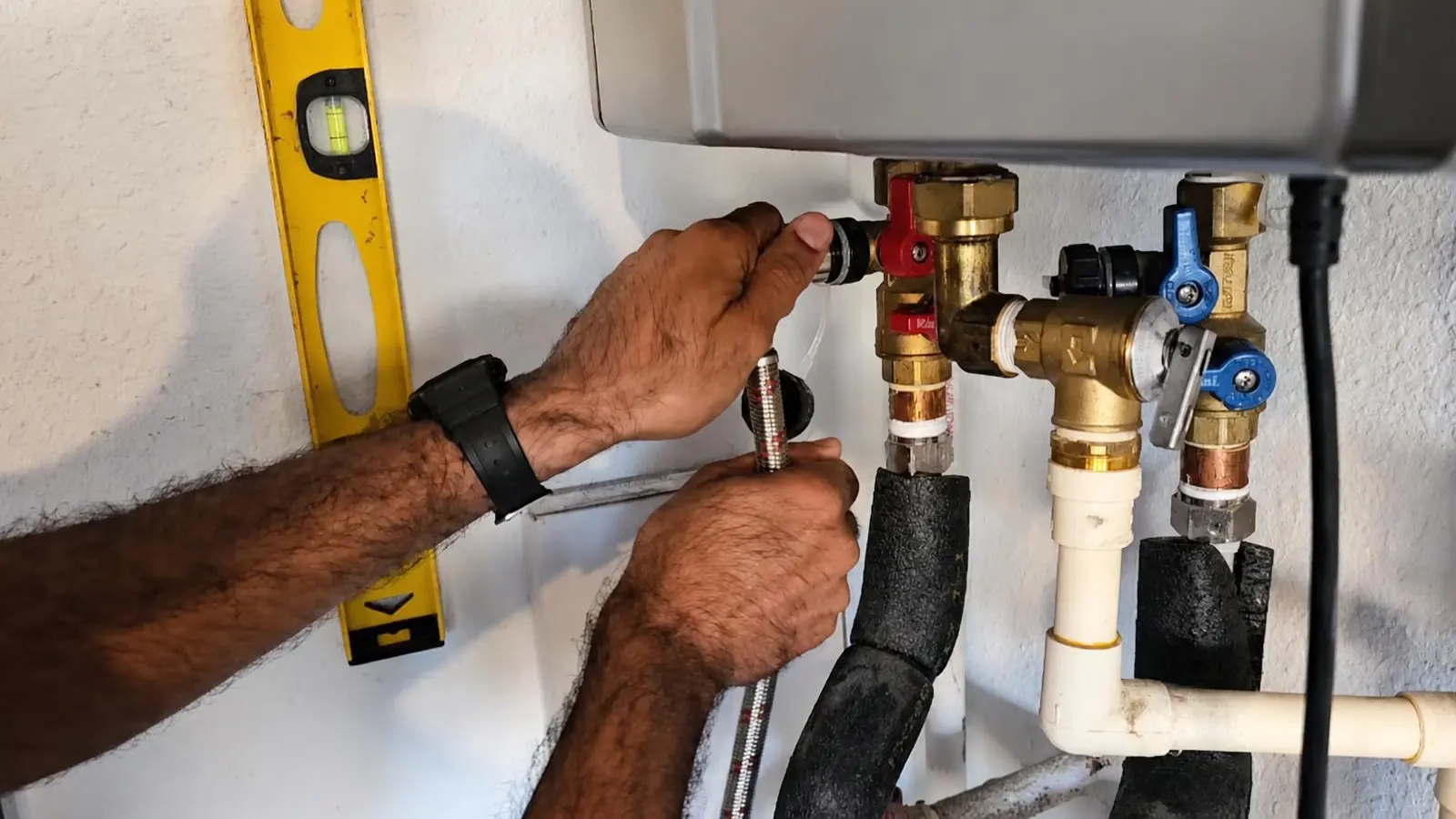 Two hands connect a hose to the brass isolation valves under a wall-mounted tankless water heater, with a yellow level on the wall beside them.