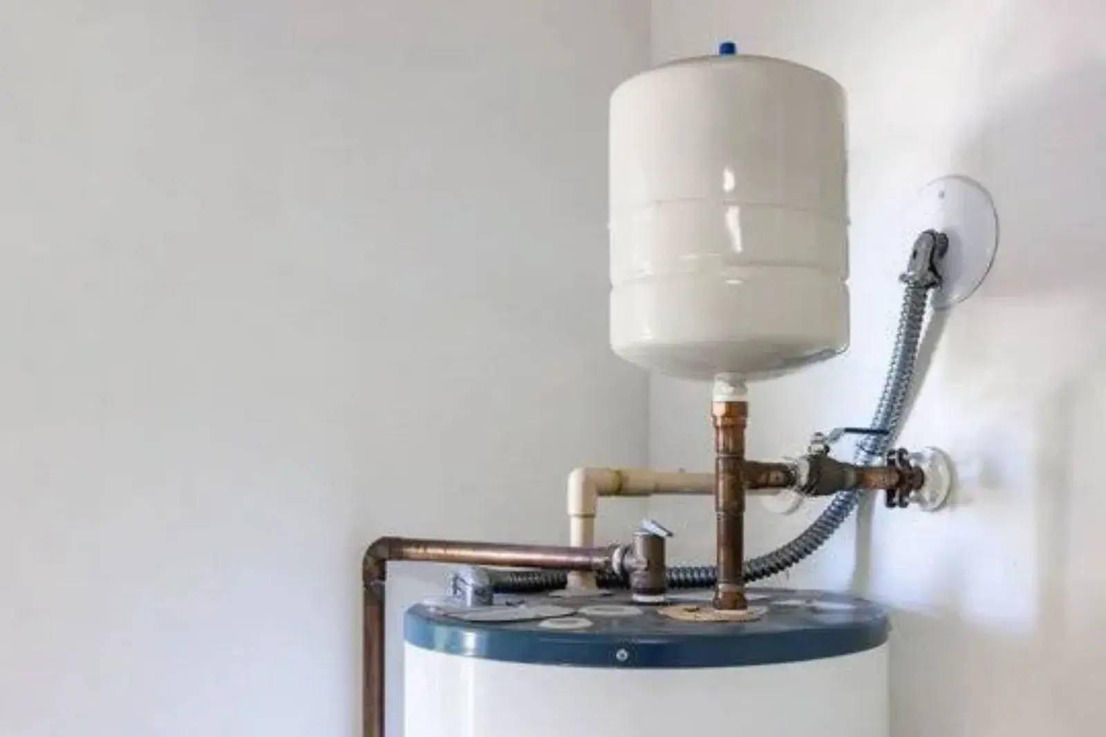 A white expansion tank mounted on a copper pipe tee above a tank-style water heater, with a flexible conduit and a relief valve nearby.